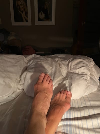 A close-up, low-angle shot shows a person's feet on a bed covered with white and blue striped sheets and a white duvet. The feet are bare and positioned near the bottom of the frame, with the toes of the left foot pointing towards the right side of the image and the toes of the right foot pointing towards the top. The lighting is dim, suggesting nighttime or a dark room, and the background is out of focus, showing a wall with two framed pictures. The image does not provide any information about the city of Oldenburg in Holstein, Germany.