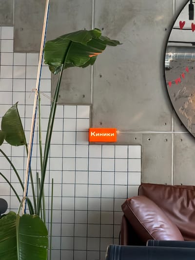 An orange illuminated sign reading "Киники" is mounted on a concrete wall. A large green leafed plant with a wooden support pole stands to the left of the sign. To the right of the sign, a round mirror with red heart and flag decorations is visible. A brown leather sofa is positioned in the lower right corner of the image. The wall features a grid pattern of white tiles. The overall scene appears to be an indoor setting, possibly a cafe or lounge.