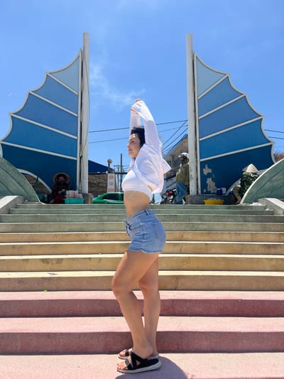 In the city of Mũi Né, Vietnam, a woman with dark hair, wearing a white crop top, denim shorts, and black sandals, stretches her arms above her head while facing right. She stands on a set of wide, outdoor concrete stairs, with some steps colored pink and others a sandy yellow. Behind her, a large, blue, sail-like structure with white accents rises against a clear blue sky dotted with faint clouds. To the left, a person in a conical hat sits on the stairs, attending to a collection of items in green baskets. Further back, a person in a green jacket and blue pants stands near a motorcycle, and another person in camouflage clothing and a conical hat is seated to the right. A green car is visible in the background, along with buildings and utility lines. The scene appears to be an outdoor public area, possibly a market or scenic overlook.