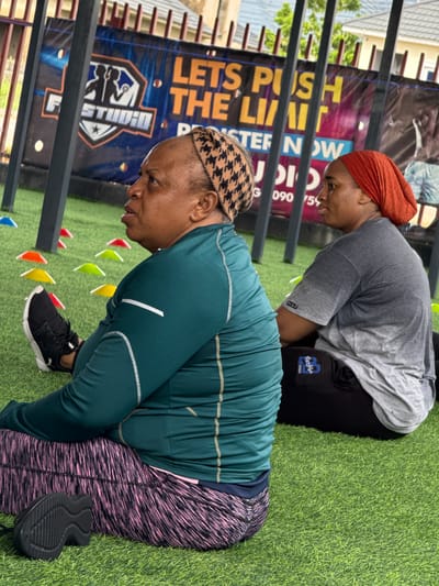 The image depicts two women sitting on a green turf surface, likely in an outdoor gym setting in Iddo Tudunwada, Nigeria. In the background, a banner reads "LETS PUSH THE LIMIT" and "REGISTER NOW." The woman in the foreground wears a green long-sleeved athletic shirt and patterned leggings, with a black sneaker visible on her left foot. The second woman, slightly behind and to the right, is wearing a grey t-shirt and black athletic pants, with an orange headscarf. Several colorful cones are scattered on the turf.