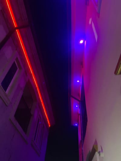 The upside-down image shows a section of a building exterior at night. A bright orange light strip highlights the edge of the building and window frames. Above, a dark wall extends to the ceiling. The ceiling has recessed lighting and blue LED strip lights. A partial view of a ceiling fan is visible. The color scheme is dominated by hues of purple, blue, and orange.