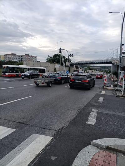 This media file captures a street scene in Prague, Czechia, during the daytime under an overcast sky. The image focuses on a multi-lane road with several vehicles, including a bus, cars, and a trailer.
In the foreground, a crosswalk is visible, with a small, grey trailer being towed by an unseen vehicle. To the left, a red and white public bus is stopped, and next to it, a dark grey SUV. Further down the road, another dark grey sedan and a black Audi are prominent. The black Audi, with license plate "1UR 4406", is facing away from the camera.
In the background, a modern pedestrian bridge spans across the road. Buildings can be seen on the left, and trees and more traffic are visible further down the road. Construction barriers and signs indicate some ongoing work or road closure. The overall mood is neutral, with no discernible emotions. The time of day appears to be late afternoon or early evening, given the cloudy conditions. No people are clearly visible in the scene.