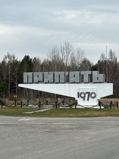 A roadside photograph of the entrance sign for Pryp'yat', Ukraine. The sign consists of large, gray, blocky Cyrillic letters spelling "Прип'ять" and the number "1970" indicating the year of the city's founding. The letters are mounted on a white, triangular pedestal. A black metal chain fence runs along the border of the sign. A paved road with markings is visible on the right side of the image. Bare trees and a cloudy sky form the background.