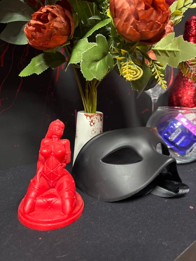A close-up, overhead shot displays a collection of items arranged on a dark, possibly black, surface with red abstract streaks. To the left, a bouquet of artificial flowers, featuring reddish-brown and orange peonies with green leaves and delicate yellow accents, is partially visible. To the right of the flowers, a clear glass jar contains a purple condom packet with white text reading "Contex classic". Adjacent to the jar is a black mask with cutouts for eyes and a red strap. At the bottom right, a bright red, sculpted figure in a dynamic pose is the most prominent object. The figure appears to be made of a waxy material and depicts a stylized human form with exaggerated features. The overall lighting is dim, suggesting an indoor setting. The presence of a condom, a mask, and the suggestive red figure could imply a private or intimate context. No specific time of day, weather, or strong emotional cues are discernible. The text "Contex classic" is visible on the condom packet.