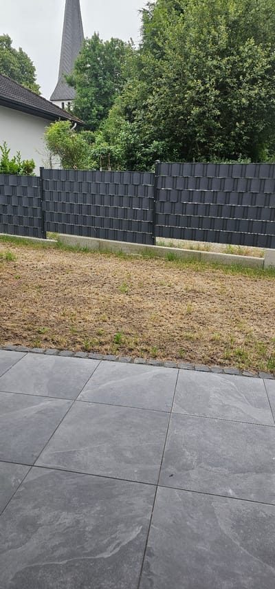 The image shows a view of a backyard in Löhne, Germany. In the foreground, there is a patio area with large grey square tiles. Beyond the patio, there is a strip of dark grey decorative edging stones separating the patio from a patch of dry, brown grass. A dark grey fence with a woven pattern runs across the middle ground, separating the grass area from the background. Behind the fence, there is a dense growth of green trees and bushes. In the upper left portion of the background, a white building with a dark roof is partially visible, and to its right, the tall, pointed spire of a church, typical of the architecture found in German towns, rises above the trees. The sky is overcast and appears to be a pale grey. There are no people or animals visible in the image.
