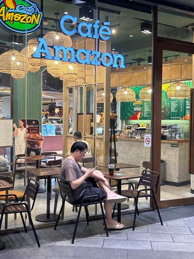 This image shows a street view of a Cafe Amazon located in Chiang Mai City Municipality, Thailand. The cafe has large glass windows, and through them, we see a man sitting at a table outside, engrossed in his phone. He is wearing a grey t-shirt and dark shorts, with one leg resting on his other knee. He is wearing light-colored sandals.
Inside the cafe, the decor features woven pendant lamps and green accents, suggesting a natural theme often associated with the "Amazon" brand. Visible signage includes "Cafe Amazon" in large blue letters reflected on the glass, and a "TASTE OF NATURE" slogan. There are also menus displayed, with one clearly showing "OPEN" and opening hours, indicating the cafe is operational. A "No Smoking" sign is also visible, a common sight in public places in Thailand. Reflections in the glass reveal more of the interior, including displays and other patrons. The lighting suggests it might be evening or overcast, as the artificial lights inside the cafe are prominent. The exterior pavement is made of light-colored stone tiles.
