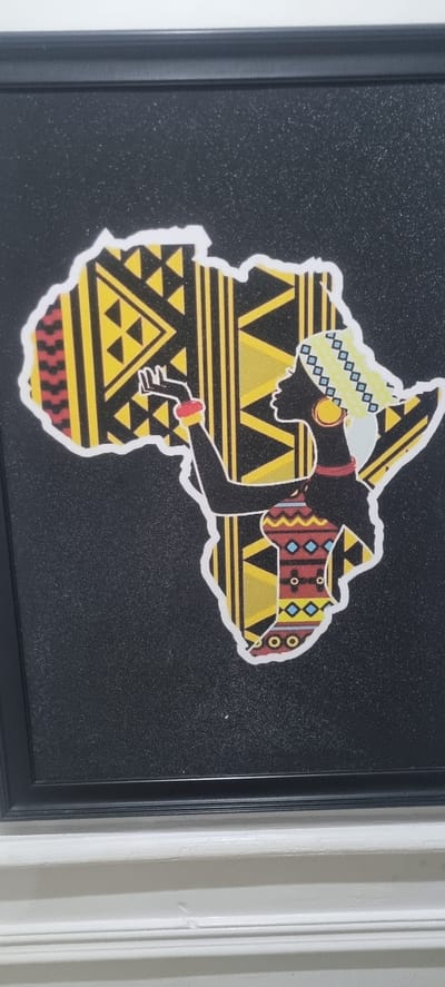 A framed artwork featuring a stylized depiction of the African continent with a woman's silhouette positioned within its borders is mounted on a wall. The artwork is characterized by a black background, over which a yellow and black patterned image of the continent is overlaid. The silhouette of the woman is black, adorned with colorful geometric patterns suggesting traditional African attire and jewelry. The artwork is framed in a black frame. The scene is located indoors, adjacent to a white wall. There is no information on the location of the city of Alasia, Nigeria.