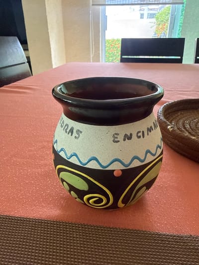 A decorative ceramic pot is centrally positioned on a vibrant coral-red tablecloth, against the backdrop of a bright room in Acapulco, Mexico. The pot features a glossy black rim, an upper section of speckled light grey ceramic with blue wavy lines, and a lower body adorned with black, green, yellow swirl patterns, and a small orange dot. The words "PIEDRAS" and "EN CIMAS" are hand-painted in black on the light grey section of the pot. To the right, part of a round, woven brown basket is visible. The tablecloth rests on a textured brown placemat or table runner. In the background, large windows reveal an outdoor scene with lush green tropical foliage and some orange-flowering bushes. Through the windows, portions of dark-colored chair backs are visible, suggesting an indoor dining or seating area. The interior features light-colored walls and large windows typical of a warm climate setting.