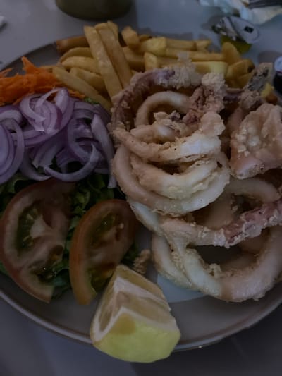 A plate of food, consisting of fried calamari rings, french fries, lettuce, tomato slices, red onion rings, and a lemon wedge, is arranged on a light gray plate. The food is located in the city of Arucas, Spain, at coordinates 28.118 latitude and -15.524 longitude. The plate is on a table, and the surrounding environment is not visible. The food appears to be a meal or snack.