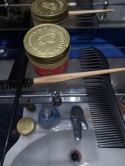 The image captures a close-up still life of various grooming tools and products, likely situated in a barber shop or a bathroom setting, with the provided location context of Tiranë, Albania. The central subjects are two golden, circular tins, each embossed with the brand "BANDIDO BARBER SHOP." The tins feature a prominent logo depicting a stylized face with a beard and mustache, surrounded by the word "BANDIDO" and small star patterns. A red label on the side of one tin is partially visible, showing the word "HAIR," indicating a hair care product.
Surrounding the tins are several grooming implements. Two black combs are visible: a long, fine-toothed comb extends horizontally across the top of the frame, and a similar comb is positioned at the bottom. Two wooden-handled brushes are also present; one has a narrow, pointed bristle head, and the bristle end of another brush is seen near the bottom edge.
The background reveals a white porcelain sink equipped with chrome faucets and a drain. A small, circular golden object, possibly another tin lid or a coin, rests near the sink's drain. A highly reflective surface, likely a mirror or polished metal shelf, creates noticeable reflections of the objects, including the backs of the combs, the other sides of the tins, and a distinct blue background element. This adds depth and a sense of order to the composition. The lighting is soft and even, highlighting the metallic sheen of the tins and the textures of the grooming tools. No activity is taking place; the image presents an organized display of items. There are no discernible cues regarding time of day, weather, or emotions within the image itself.