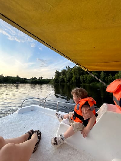 The image captures a serene scene on a body of water, likely a river or lake, during what appears to be late afternoon or early evening, given the soft, warm lighting. A small boat, possibly a paddle boat given the white textured deck and lack of visible motor, is the primary setting. A young child, wearing an orange life vest over a tan t-shirt and beige shorts, is seated on the edge of the boat, looking out towards the water. Their feet, clad in white socks and sandals, rest on the textured deck. In the foreground, the legs of an adult are visible, wearing brown sandals. The boat's interior has white molded seating. Beyond the boat, the water is calm, reflecting the sky and the lush green treeline that borders the opposite bank. The sky is a blend of soft blue with scattered wispy clouds, illuminated by the warm glow of the setting sun. The overall impression is one of peaceful recreation in a natural environment. The location is specified as Klin, Russia, suggesting this is a local waterway within or near the city.