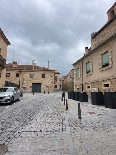 The image shows a cobblestone street in Segovia, Spain (latitude: 40.952, longitude: -4.123) with a car parked on the right. The street runs uphill to the left, where the perspective is distorted due to the angle of the image. There are stone buildings on either side of the street, which appear old and weathered. The sky is overcast with clouds. The buildings along the street have windows and doors. The cobblestone street is worn and uneven. The car is a silver sedan. There are metal poles and other objects lining the sides of the street.