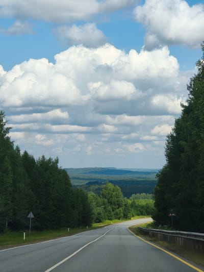 The image depicts a road curving through a dense forest with a vast, rolling landscape of trees in the distance. The sky is filled with dramatic, fluffy white clouds against a clear blue background. The road itself is asphalt, with white lane markings and a yellow line delineating the edge. Guardrails and road signs are visible along the side of the road. The scene appears to be a scenic drive, likely in a rural or natural area. The specific location in the city of Krasnye Gorki, Russia, implies a natural setting within or near that locale, consistent with the imagery. There are no people or animals visible in the frame.