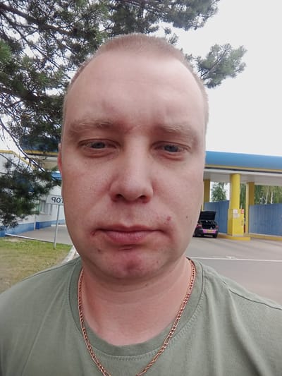 The media file is a selfie of a fair-skinned person, likely male, with short blonde hair and blue eyes. He is wearing a green t-shirt and a thin gold chain. The image is oriented sideways, making it difficult to discern the full context of the scene.
The person's face takes up a significant portion of the frame, with his gaze directed towards the camera. His expression is neutral, though a small red mark is visible on his left cheek. The background appears to be an outdoor setting with some trees and a structure that looks like a gas station canopy with blue and yellow supports. There are some visible details of a car in the background.
Based on the text "1.3" visible on a yellow sign, and the "PRO" lettering on the side of the car, it could suggest a location related to services or a particular area. The presence of trees and the overcast sky suggest it might be a cloudy day. The overall impression is a candid snapshot taken outdoors.
The context provided places the location as Mahilyow, Belarus, which could indicate that the text visible in the image is in Belarusian or Russian.