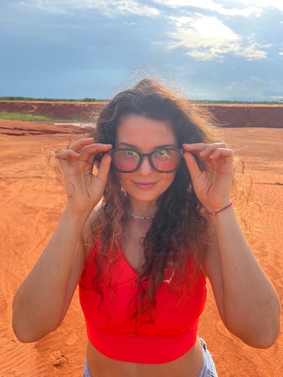 In a medium shot, a woman with curly brown hair stands outdoors in a sandy, arid environment. She is wearing a bright red sports bra and jeans, and she is adjusting black-rimmed glasses. The background consists of a vast expanse of orange sand, with a distant red earth cliff and a sky with scattered clouds. The lighting suggests it is late afternoon. The location is identified as Mui Ne, Vietnam, a coastal resort town known for its sand dunes.