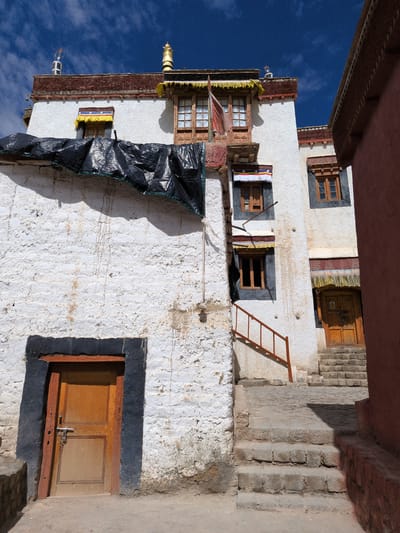 The image captures a view of a traditional Tibetan architectural style building with white walls and wooden accents, set against a bright blue sky with scattered clouds. The building features multiple windows with wooden frames, some adorned with colorful fabric awnings. A large black tarp is draped over a section of the building's upper exterior. There are stone steps leading up to a wooden door on the right side of the image. A metal railing is visible along a set of stairs on the left side. The ground is a mix of dirt and stone paving. The overall scene appears to be a courtyard or alleyway within a monastery or ancient settlement. The location is identified as Choma, India. There are no people visible in the image.