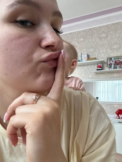 The image shows a close-up of a woman's face, with her finger to her lips in a shushing gesture. She is wearing a light-colored top and a ring on her finger. In the background, a toddler is visible, wearing a pink shirt. There are shelves in the background with books and framed pictures. The scene appears to be indoors, possibly a home. The provided information states the location is the city of Rossosh, Russia.