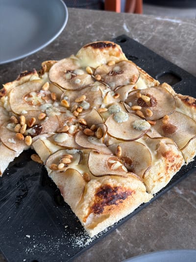 A close-up overhead shot shows a pear and pine nut flatbread pizza on a black cutting board. The pizza is sliced and arranged on the board with some of its crust slightly burnt. The flatbread is topped with thin slices of pear, pine nuts, and small crumbles of blue cheese, all covered with melted cheese. A portion of a gray plate is visible in the upper left corner, and the surface beneath the cutting board appears to be a marble countertop. The scene is set in a restaurant in Bishkek, Kyrgyzstan.