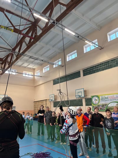 The image shows an indoor climbing or adventure course facility in Berezniki, Russia. Several people are wearing helmets and harnesses, suggesting they are participating in the course. Some individuals are on ropes or cables, while others stand on the ground observing. The environment consists of a large, indoor space with a climbing structure, nets, and ropes. There are windows along the upper walls of the building. A basketball hoop is present, suggesting the location is a multi-purpose gymnasium or recreational facility. There is a banner visible in the image with text in Russian.