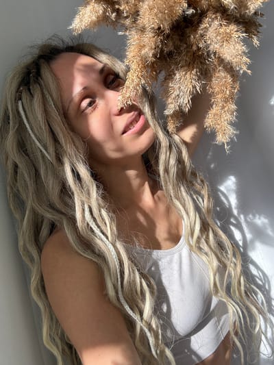 An overhead medium shot captures a person with long, wavy blonde hair lying on a light-colored, possibly white, surface. The individual is positioned diagonally across the frame, with their head towards the upper left. They are wearing a simple white ribbed tank top. Their eyes are partially closed or looking downwards, and their lips are slightly parted, suggesting a relaxed or contemplative state.
A prominent bunch of dried, light brown ornamental grass or reeds, likely pampas grass, occupies the upper left quadrant of the image, partially covering the person's face on that side. The person's long, flowing blonde hair, which appears to have integrated thin white braided strands, cascades around their head and shoulders, spreading across the surface.
The scene is illuminated by strong, direct sunlight, casting distinct, sharp shadows across the surface and the person's form. These shadows, some resembling abstract organic shapes or foliage, create a high-contrast visual effect. The lighting emphasizes the textures of the hair, the dried grass, and the ribbed fabric of the tank top. No visible text is present in the image. The location context for the image is Rusanovo, Russia.