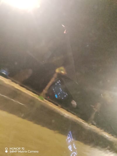 A blurry, poorly lit photo shows a smartphone resting on a surface with a white substance. The phone's screen is illuminated, displaying what appears to be text and icons. Above the phone, a reflection of a light source is visible, further obscuring the details. The overall image is murky and lacks clarity, with limited discernible features beyond the smartphone and light reflection. The lower portion of the image includes the text "HONOR 70" and "54MP Matrix Camera".