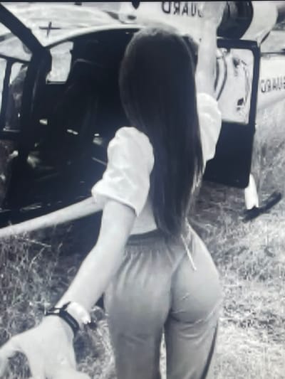 In a grayscale image, a person with long dark hair is captured from behind, standing near an open helicopter door. The person is wearing a light-colored, puffy-sleeved top and loose-fitting pants, with a watch on their left wrist. The helicopter appears to have the word "GUARD" visible on its side, suggesting it may be a coast guard or rescue helicopter. The background shows a grassy, outdoor environment, possibly near a body of water, though this is not explicitly clear. The image does not provide any specific information or visual cues to determine the location within the city of Shakhovskaya, Russia.
