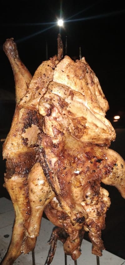 A grilled chicken hangs vertically on metal rods under dim lighting. The skin is browned and glistening with rendered fat, showing areas of darker charring. One leg is prominently displayed on the left. Above, a bright light, possibly a streetlight, shines through the darkness.