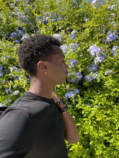 A young Black person with dark, curly hair lies on their side amongst a lush green bush with small blue flowers. The person is wearing a black t-shirt with a ribbed texture on the sleeve and chest. They have a silver hoop earring in their left ear and a brown leather bracelet with silver studs on their right wrist. Their eyes are closed, and their head is tilted back, suggesting a moment of relaxation or contemplation. The bright sunlight indicates it is daytime. The foliage and flowers suggest a natural, outdoor setting, likely a garden or park. The location context of Sant Adrià de Besòs, Spain, places this scene in a Mediterranean environment. The overall mood is peaceful and serene. There is no visible text in the image.