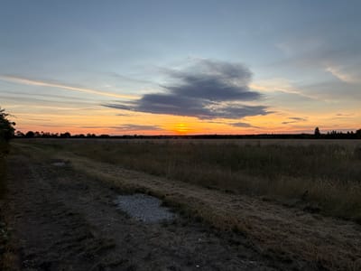 The image captures a rural landscape at dusk in East Hertfordshire, United Kingdom. A dirt path runs diagonally from the lower left corner towards the center of the frame, bordered by dry, mown grass. Beyond the path, a field of tall, dry grass stretches to the horizon. In the distance, a line of trees and silhouetted structures mark the edge of the field. The sky is a gradient of colors, transitioning from a pale blue at the top to warm oranges and yellows near the horizon, indicative of a sunset. A few scattered clouds, some dark gray and others wispy and light, are present in the sky. The overall impression is one of a peaceful, natural setting during twilight.