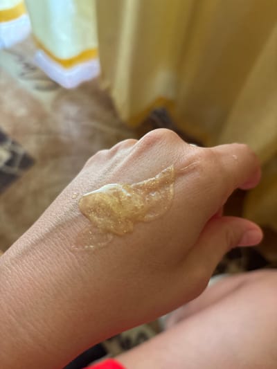 A close-up shot shows a person's hand with a dollop of shimmering, golden gel on the back. The gel appears somewhat viscous and contains fine glitter particles. The skin of the hand is visible with texture and pores. The background is softly blurred, showing elements of an indoor environment with what appears to be yellow curtains and a patterned surface, possibly a rug or blanket. The image does not contain any elements that would allow for identification of a specific location, including Krasnaya Polyana, Russia.