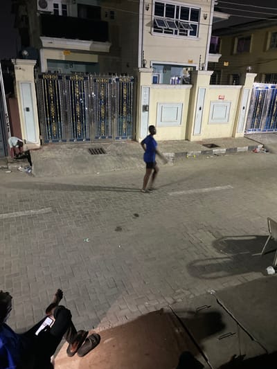 An image captured at night depicts a street scene. A person in a blue shirt and dark shorts appears to be running or walking quickly. Adjacent to the street are buildings with decorative gates. At the lower right of the frame, another person is visible, seated and using a smartphone. The street is paved with bricks or tiles. Shadows are cast from an unseen light source.