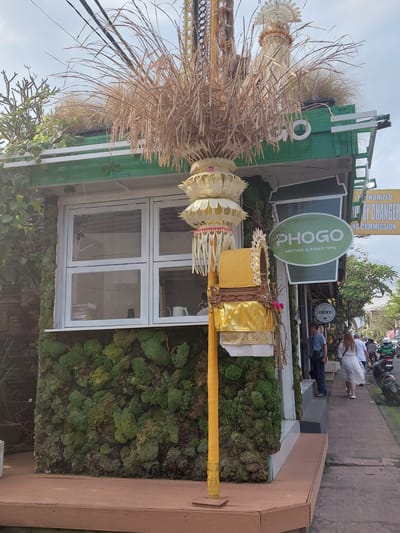 In Ubud District, Indonesia, on a cloudy day, a building facade is adorned with vibrant green moss and dried grass decorations. A tall, yellow pole with ornate, layered ornaments sits in the foreground. A white-framed window is visible on the moss-covered wall. To the right, a green sign with white text reads "PHOGO ANYTIME IS PHOGO TIME." Further down the street, people walk along a paved sidewalk. A motorcycle is parked nearby, and other vehicles are visible on the road. The scene suggests a commercial area, possibly a cafe or shop, with a unique, nature-inspired aesthetic.