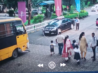 The image depicts a scene in Bengaluru, India, likely near a school entrance. A yellow school bus is parked on the left side of the frame. A black sedan is parked in the middle of the frame, behind a white picket fence. Several people are visible, including adults and children in school uniforms, suggesting drop-off or pick-up time. Some people are walking towards or away from the camera. The environment includes trees, banners, and a building with a blue awning in the background.