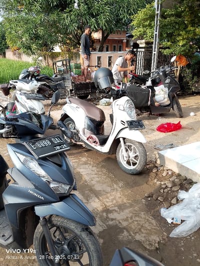 The image shows a street scene in Kecamatan Haurgeulis, Indonesia, at coordinates -6.431 latitude and 107.943 longitude. There are five motorcycles parked on a concrete street. The motorcycles are facing different directions. A blue Honda motorcycle is parked on the lower left side of the image. Behind it is a white motorcycle with the license plate E 6898 QAL 18-28. A white motorcycle with the license plate F 8127 KY is parked on the upper right side of the image. A white motorcycle with the license plate E 5745 RAI is parked on the left of the image. Another motorcycle is in the background, behind the motorcycles. Two people can be seen in the image: a man in a black shirt and gray pants standing on a raised platform at the upper left side of the image, and a man in a red shirt and gray pants standing on a platform beside a motorcycle. The background shows a house and green trees. The image is taken on a sunny day, and the street is covered with gravel and debris. A white plastic bag is lying on the street, and a white bag is lying on the ground beside the motorcycle on the right side of the image. The image was captured on a Vivo Y19s phone on February 28, 2023, at 07:33. The image was captured by Akhtar and Arvan.