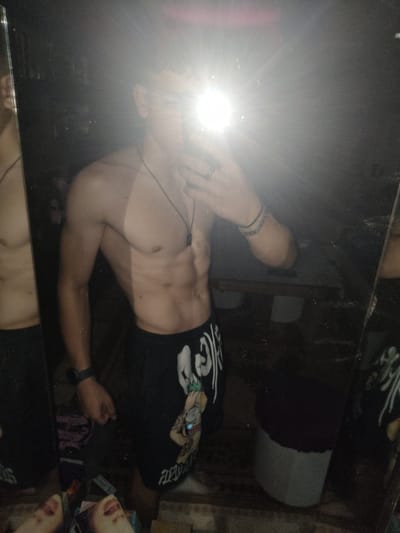 This image, captured in Yambol, Bulgaria, depicts a young male taking a mirror selfie in a dimly lit indoor setting. The subject is shirtless, showcasing a lean physique with visible abdominal musculature. A distinct tan line is present across his upper chest and shoulders. He wears a dark necklace with a rectangular pendant. His face is obscured by the bright flash from the smartphone he holds in his right hand. A ring is visible on the ring finger of his right hand. His left wrist is adorned with multiple dark bracelets and a dark watch. He wears dark shorts featuring white graphic designs, including a cartoon character with a crown and visible text "R1PND" on the viewer's left leg. The background, reflected in the mirror, reveals a dark, indistinct room. A white or light grey bucket, topped with dark fabric, is positioned on the floor to the subject's right. Reflections of what appear to be papers or magazines are visible on the floor at the bottom left of the mirror.
