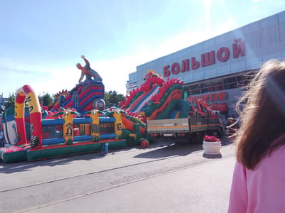 The image depicts an outdoor scene in Zavodoukovsk, Russia, featuring a large inflatable playground with characters such as Spiderman and a dragon. To the right, a building with the sign "БОЛЬШОЙ" (Bolshe - Big) is visible, indicating it's likely a shopping center or department store. In front of the building, a truck with a crane is parked, and a white flower pot filled with red flowers sits near the truck. A person, partially visible and wearing a pink sweatshirt, is in the foreground, facing away from the camera. The sky is clear and blue, suggesting a sunny day. The overall atmosphere suggests a public event or a day out for families.