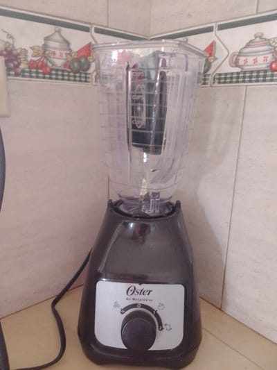 In La Tagua, Venezuela, an Oster All-Metal Drive blender sits on a countertop against a tiled backsplash. The blender is black with a clear plastic pitcher. The pitcher has measurement markings visible. The control panel on the blender's base displays various speed settings indicated by icons. To the left of the blender, a black power cord is visible. The backsplash features a decorative border with images of fruit and pottery.