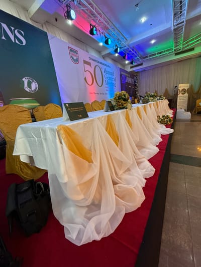 The image depicts a banquet setting inside an event hall. A long table is draped with white fabric with gold accents and decorated with floral arrangements. The floor is covered with a red carpet. Behind the table is a large banner announcing the "50th Annual General Meeting" of "Champion Breweries PLC," with the tagline "Nourishment of Champions" and the date "Thursday, 21st May, 2026." The banner also mentions "Scaling Growth, Profit & Productivity Sustainably." A nameplate on the table reads "Mr. Thompson Owoka, Non-Executive Director." Stage lighting is visible in the upper-left corner of the image, and a sign proclaiming "Brewing Joy, Enriching Champions" is further down the table on the right. The sign includes the location: Itirin, Nigeria. A black camera case sits on the floor beside the table.