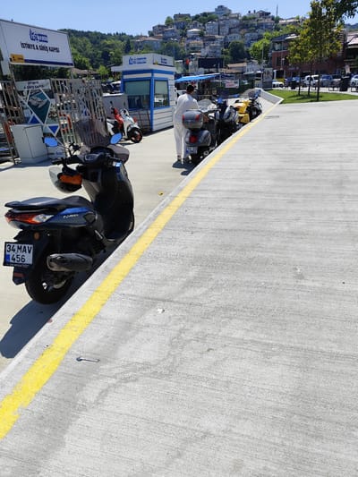In Sarıyer, Turkey, a sunny daytime scene unfolds at a marina entrance. A blue Yamaha NMAX scooter, bearing a Turkish license plate "34 MAV 456", is parked on a concrete pathway marked with a bright yellow line. Behind it, a line of other scooters, including a yellow one, are also parked. A person in white attire stands near the scooters, facing away from the camera, appearing to be an employee or attendant. Further in the background, a white and blue booth labeled "İSTİNYE MARIN" suggests a ferry or boat service terminal. Beyond the terminal, a dense collection of houses with red-tiled roofs cascades down a lush green hillside under a clear blue sky, indicating the urban and hilly geography of the area. Trees are scattered throughout the background. The overall impression is a bustling waterfront with transportation services.