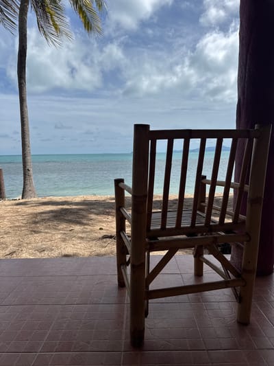 An eye-level, full shot captures a simple bamboo chair on a tiled porch overlooking a beach in Baan Tai, Thailand. To the left of the chair, a tall palm tree stands with its fronds reaching towards a cloudy sky. Beyond the tree and the chair, a sandy beach leads to the ocean, which is a tranquil turquoise. The water meets the horizon where a faint outline of land is visible under the pale sky. The foreground is dominated by the textured, reddish-brown tiles of the porch, with the bamboo chair placed in the center. The chair's slatted back and seat are visible, and its legs rest on the tiles. A dark, textured pillar is to the right of the frame, partially obscuring the view of the chair. The overall scene conveys a serene, tropical atmosphere.
