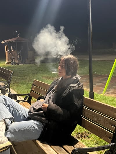 The image captures a nighttime scene in what appears to be a park or outdoor public area in Lozarevo, Bulgaria. A young individual with curly hair is seated on a wooden park bench, wearing a dark jacket or hoodie, light blue jeans, and light-colored sneakers with patterned socks.

The primary activity is smoking; the person holds a lit cigarette and exhales a significant plume of white smoke that billows prominently to the left, catching the ambient light. Their expression is partially obscured by their hair and the angle, but they appear to be looking downwards or to the side.

The setting is dimly lit by artificial light sources. A black lamppost stands vertically in the center, and a bright, greenish-yellow horizontal strip of light is visible in the upper right background. Lush green grass surrounds the benches, and a paved or tiled path is also visible. In the distant background, to the lower left, a gazebo-like wooden structure is faintly discernible, also illuminated by some light. The benches feature ornate black metal frames with a decorative grape or vine pattern, supporting wooden slats. The overall atmosphere is quiet and somewhat contemplative, characteristic of an evening in a public space. No discernible text is visible in the image.