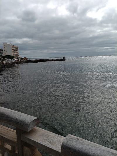 This image presents a coastal view in Agüimes, Spain, under an overcast sky. In the foreground, a weathered, light-colored wooden railing with a horizontal top rail and a lower support extends across the bottom of the frame, partially obscuring the immediate water below. The midground on the left features a multi-story building with a light-colored facade and multiple balconies, positioned along the shoreline. Adjacent to this building, a long, dark-colored concrete or stone pier/breakwater extends into the water from the coast towards the right, ending with a visible structure. The predominant feature of the midground and background is the dark grey ocean water, displaying numerous small ripples and reflections of the cloudy sky. The sky is entirely covered with thick, grey clouds, exhibiting some brighter patches. No individuals, explicit actions, or interactions are visible within the scene.