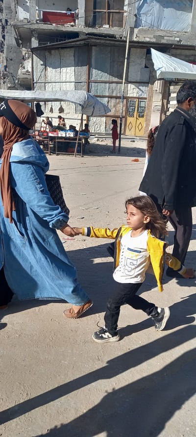 In Khan Yunis, Palestinian Territories, a young child with long, dark hair runs forward, holding the hand of an adult woman who is wearing a blue denim dress and a brown hijab. The child wears a yellow jacket over a white t-shirt with text and graphics, and black pants. The adult is walking away from the camera, and the child is walking next to them, looking to the right. In the background, there is a partially destroyed building with visible damage. Several other people are present in the background, including an adult man in a dark coat. A small market stall with various items is also visible. The ground is a dirt and sand surface, with shadows cast by structures and people.
