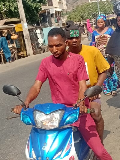 A man in a red shirt drives a blue and white motorcycle. Another man, wearing a yellow shirt and a black baseball cap, sits behind him. A woman in a patterned blue top sits behind the second man, facing forward. They are on a paved road, with buildings and other people visible in the background. A streetlight stands beside a building to the left of the frame.
