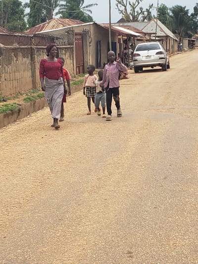 A group of people, including a woman and three children, walk along a dirt road in Kachia, Nigeria. The woman, in the foreground, wears a red shirt and a patterned skirt. She is walking to the left of the frame. Behind her, a child in a red shirt walks. To the right of the woman, two boys walk ahead. The boy in front wears a pink shirt and dark pants. The boy behind him wears a light-colored shirt and patterned shorts. The road is flanked by buildings with corrugated metal roofs and appears to be a residential area. A silver car is parked on the right side of the road, further down. The sky is overcast.