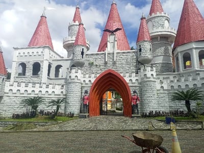 The image displays a multi-turreted castle-like building in Cipanas, Indonesia, constructed with white faux-stone block walls and battlements. Numerous conical roofs, predominantly red, cap the turrets; a white flag flies from the tallest turret on the left, and a black bird sculpture adorns a central turret. The main entrance features a prominent reddish-orange arched gateway, within which a dark wrought-iron gate is visible. Two life-sized statues, depicting figures in red British Royal Guard uniforms and black shako hats, stand on either side of the archway. The foreground consists of a dark, irregularly patterned paved forecourt. To the right, a red-framed wheelbarrow filled with dark material and a yellow and white striped traffic cone with a blue object attached to its top are present. Green garden beds with small palm-like plants are situated along the base of the castle walls. The sky above is partially cloudy blue.
