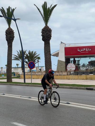 A cyclist wearing a red helmet and dark clothing rides a white Canyon bicycle on a paved road. The location is identifiable as M'Diq, Morocco, due to the presence of a building with Arabic script reading "Shwarma Avenue" and "حارة الشورما". Palm trees line the street. A blue and red no-parking sign is visible in the background. The sky is overcast.