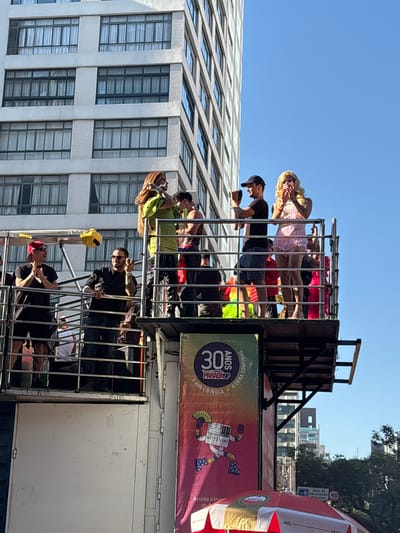 This image captures a vibrant scene from what appears to be the 30th São Paulo LGBT Pride Parade ("Parada SP") in São Paulo, Brazil, likely on Paulista Avenue given the urban setting with tall buildings. The scene focuses on a multi-level parade float under a clear blue sky.

On the upper level of the float, several individuals are visible. On the far left, a man in a black t-shirt, red baseball cap, and sunglasses is clapping. To his right, another man in a black t-shirt and sunglasses holds a mobile device, possibly recording, and is waving. In the center, a woman with long, light brown hair, wearing a bright green top and dark, possibly leather, pants and boots, poses with her arm raised. Next to her, a man with dark hair, shirtless or wearing a very light tank top, looks towards her. On the far right, a person with a long blonde wig, heavy makeup, and a frilly pink minidress is posing dramatically with hands near their face. A yellow cap or hat is hanging from the railing between the clapping man and the man with the phone.

The lower section of the float features a large vertical banner. The top of the banner displays "30 ANOS PARADA SP" in white and purple text. Below, it reads "A RUA CONVOCA A URNA CONFIRMA" (The street summons, the ballot box confirms). A stylized cartoon character named "VOTINHO," depicted as a computer screen with arms and legs wearing star-spangled boots and holding a fan, is central to the banner design. The "Amstel" beer logo is visible at the bottom of the banner, which is predominantly orange, purple, and pink.

Below the float, a red and white striped umbrella with the "Amstel" logo is partially visible, suggesting a vendor or public seating area. To the right of the umbrella, a blue street sign with white text, partially obscured, indicates "AREA DE VELOCIDADE REDUZIDA" (Reduced Speed Area) and a speed limit of "40". Green foliage from trees is visible above the sign and to the right.

In the background, a large, multi-story white building with numerous windows, each fitted with white curtains, dominates the left side of the frame. Other buildings are visible in the distant background on the right, under the bright sunlight. The overall impression is one of a public celebration or protest in a dense urban environment.