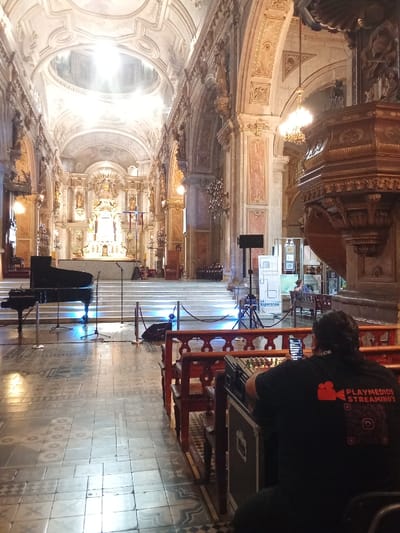 Inside an ornate church, a sound engineer works at a mixing console. A black grand piano and microphones are set up on a raised platform. The church's interior is lavishly decorated with intricate details and a bright dome. A banner displays the word "Esperanza." The engineer wears a black t-shirt with "PLAYMEDIOS STREAMING'S" printed on the back. The floor is tiled in a geometric pattern.