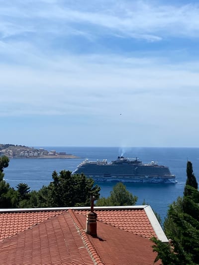 In the city of Šušanj, Montenegro, on a clear day, a large cruise ship sails on the calm, blue sea. The ship, which appears to be the Norwegian Encore, has a distinctive blue and white hull. Smoke billows from its stacks. In the distance, a landmass with industrial structures, possibly storage tanks, is visible on the left. In the foreground, the terracotta tiled roof of a building, complete with a brick chimney, occupies the lower portion of the frame. Lush green trees partially obscure the view of the sea and the ship. The sky above is a bright blue with scattered white clouds. A small, dark object, possibly a bird or a drone, is visible in the sky above the cruise ship.